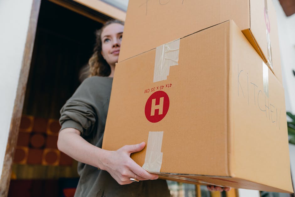 A woman with curly brown hair, wearing a grey long-sleeved top, is inside a property holding a large cardboard moving box labeled with a red 'H' and tape sealing the sides, as part of a home relocatio