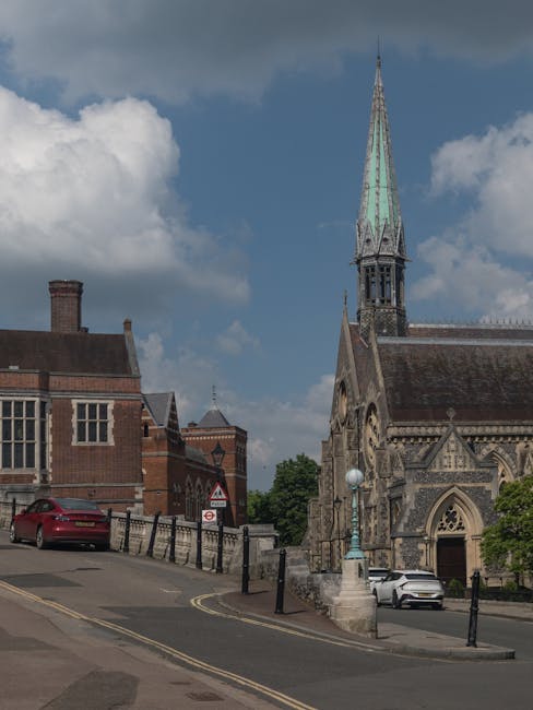 A street scene featuring a historic church with a tall, narrow spire topped with a weather vane, located on a slight incline. Nearby, there are traditional brick buildings, including one with a chimne